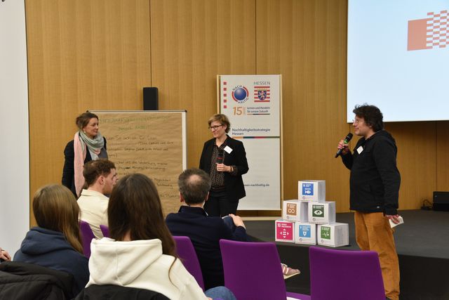 3 Personen, die vor einem Publikum auf der Bühne stehen