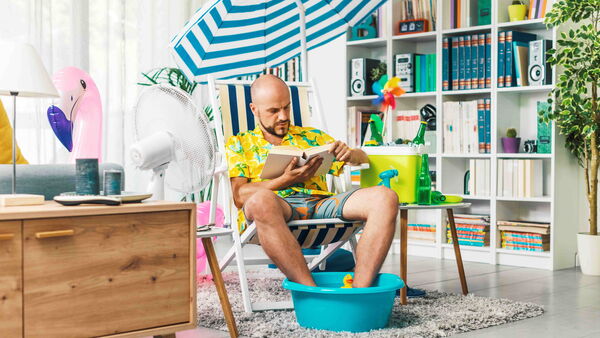 Mann verbringt seine Ferien zu Hause und liest ein Buch, dabei stehen seine Füße in einer Schüssel mit Wasser.