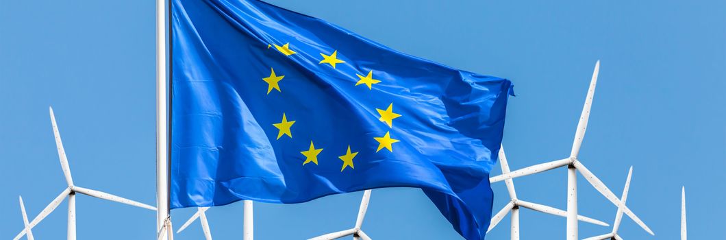Eine EU-Flagge im Vordergrund weht vor einigen Windrädern und blauem Himmel.